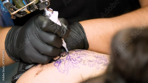 close-up, in a tattoo salon , a specialist is doing a tattoo on woman's back, black paint floral ornament. a man works in special black gloves, on special equipment. photo