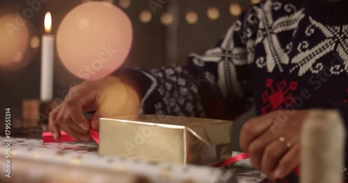 Handsome dark skinned man in black white and red fairisle sweather wrapping Christmas present among decorations and lights