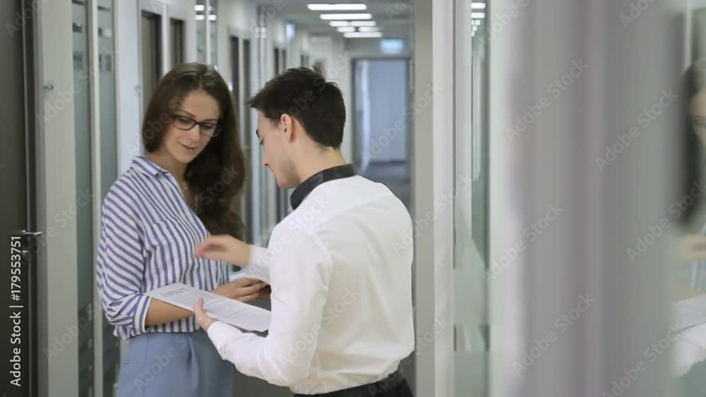 Man and woman are talking, holding tablet and documents, standing in company. Beautiful brunette in glasses turns over white sheets and gives colleague, with charming smile, guy holds silver gadget in Man and woman are talking, holding tablet and documents, standing in company. Beautiful brunette in glasses turns over white sheets and gives colleague, with charming smile, guy holds silver gadget in