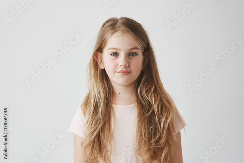 Close up portrait of good-looking young girl with blond hair in pink dress, looking in camera with calm expression, posing for photoshoot in school.