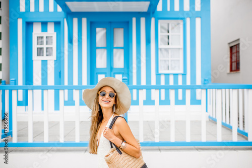 Young woman in swimsuit and sun hat standing with old traditional blue striped house on the background in Costa Nova resort in Portugal