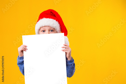 Cute little girl in Santa Claus hat with a white blank paper sheet. Christmas, x-mas, New Year, Winter, people, advertisement, sale concept. © nazarovsergey
