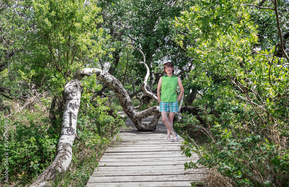 horizontal image of a caucasian woman posing on a wooden plank walk way surrounded by dense lush green trees on a warm summer day.
