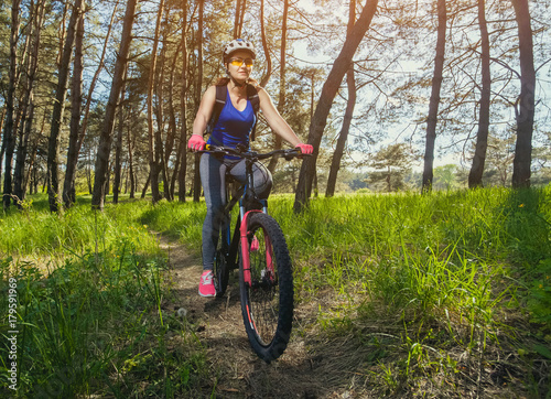 One young woman - an athlete in a helmet riding a mountain bike outside the city, on the road in a pine forest on a summer day.