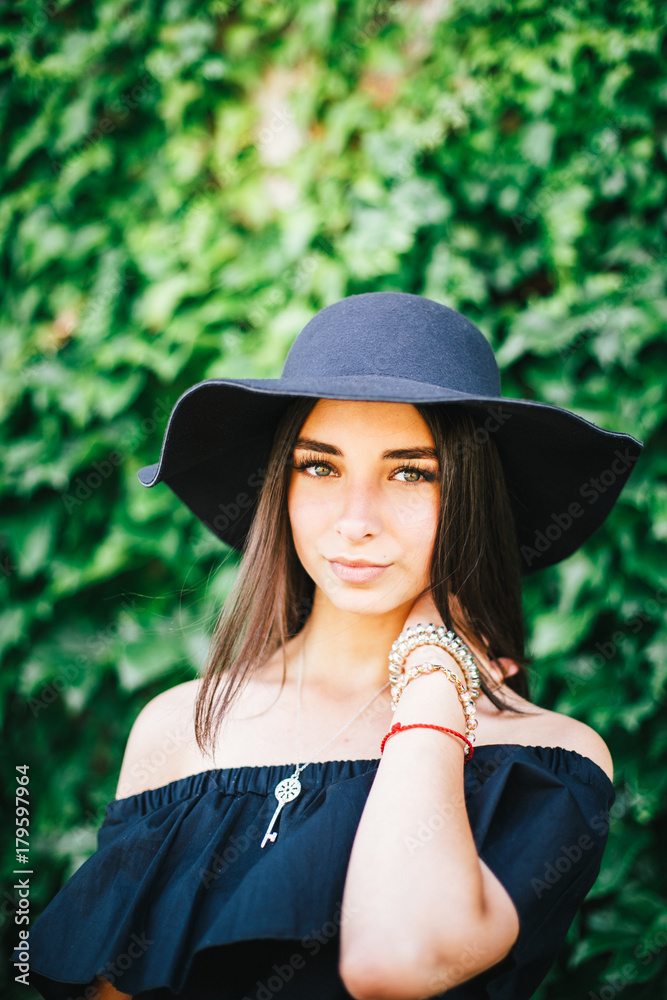 beautiful young sexy girl with swarthy skin and brunette with black hair dressed in a stylish black dress and hat on background of green foliage