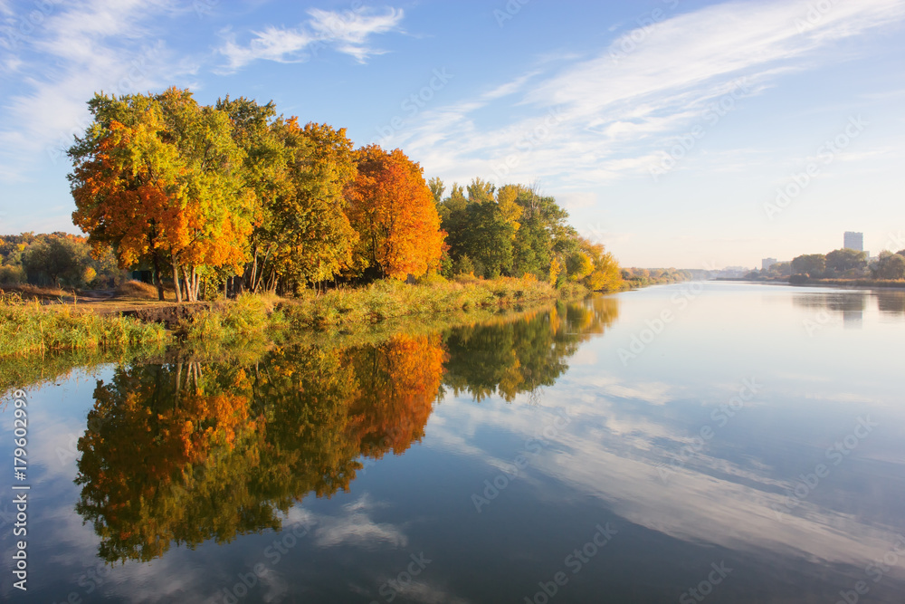 Autumn landscape in city park. Orange trees on river bank reflected in water. Beautiful blue cloudy sky