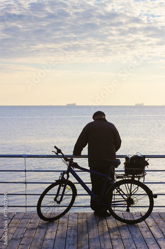 Man with bicycle. Autumn morning seascape with ships on horizon. Silhouette of elderly man on wooden pier. Pastel blue colors. © arvitalya