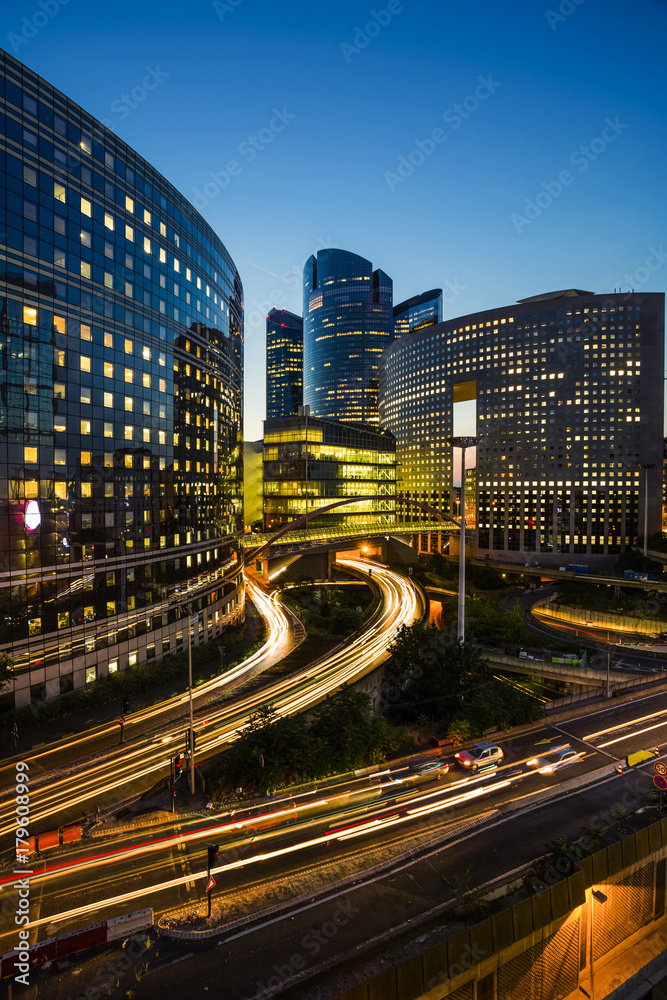 Skyscrapers in Paris business district La Defense. European night cityscape with dynamic street traffic, car lights and glass facades of modern buildings. Economy, finances, transport concept. Toned