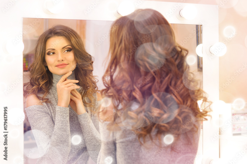 Fototapeta premium Beautiful sexy woman touching her face and looking at her reflection in the mirror.Woman with beautiful makeup and hair in curls.