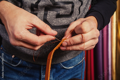 A close-up of a weed man in a T-shirt and jeans makes a genuine leather strap, makes holes for a buckle for buckles, in background is natural leather