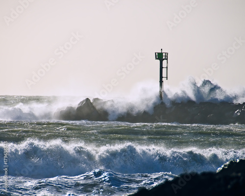Tower on rock jetty with wave crashing over it during sunset.