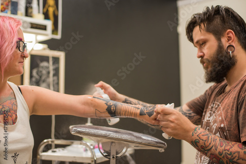 A bearded tattoo artist , in a brown t-shirt fills the hand tattoo the girl with the pink hair and tattoos and piercings all over her body photo