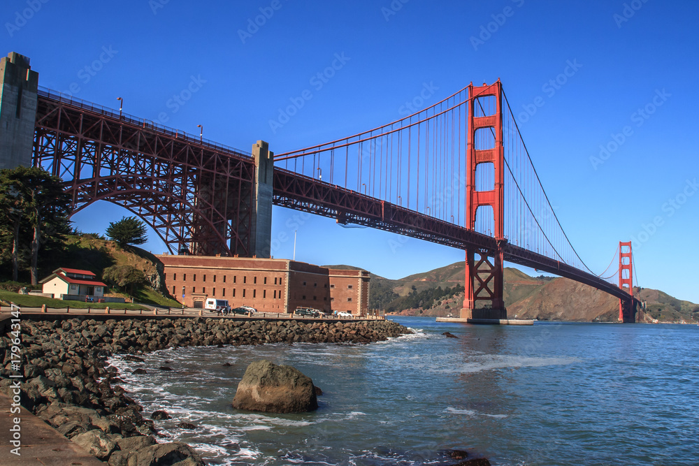 Fototapeta premium Golden Gate bridge from the San Francisco side. View of Fort Mason on the left and rocks in the foreground. A blue sky is in the background. It is a horizontal image.