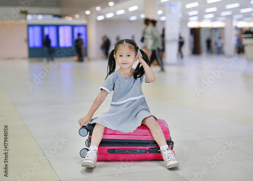 Happy little asian child girl sitting on suitcase at the airport. Waiting for flight.