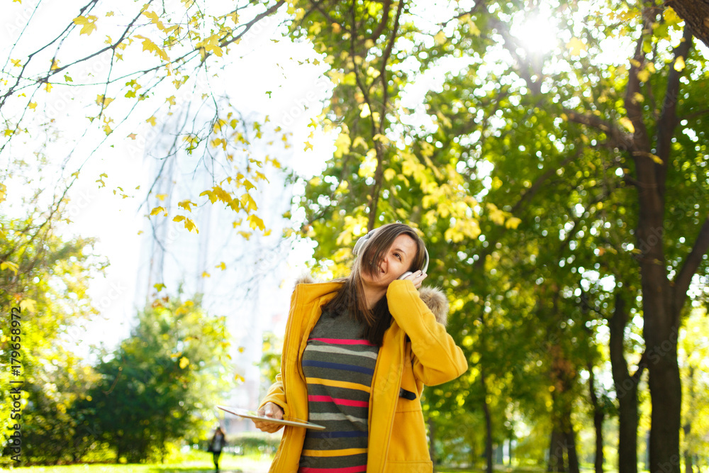 A beautiful happy cheerful brown-haired woman in a yellow coat and striped longsleeve rejoices with a tablet in her hands and white headphones in fall city park on a warm day. Autumn golden leaves. A beautiful happy cheerful brown-haired woman in a yellow coat and striped longsleeve rejoices with a tablet in her hands and white headphones in fall city park on a warm day. Autumn golden leaves.