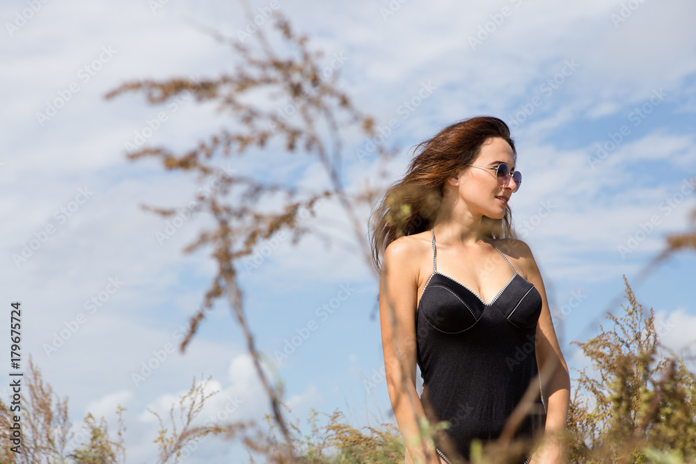 Portrait of a young sexy girl standing on a background of beach, sand and sea. She wearing black swimwear, blue shirt and sunglasses Portrait of a young sexy girl standing on a background of beach, sand and sea. She wearing black swimwear, blue shirt and sunglasses
