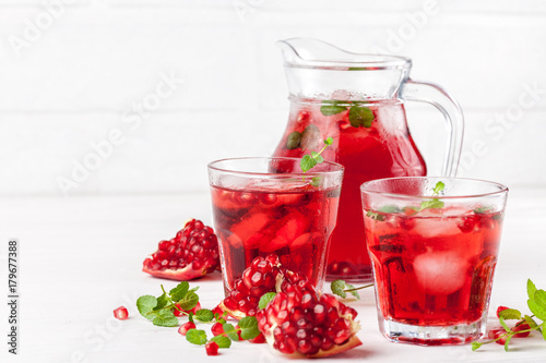 Pomegranate cocktail with ice and mint in beautiful glasses and jug, fresh ripe pomegranate on white wooden background. Sweet red juice. Close up photography. Selective focus