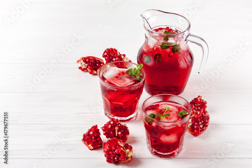 Pomegranate cocktail with ice and mint in beautiful glasses and jug, fresh ripe pomegranate on white wooden background. Sweet red juice. Close up photography. Selective focus