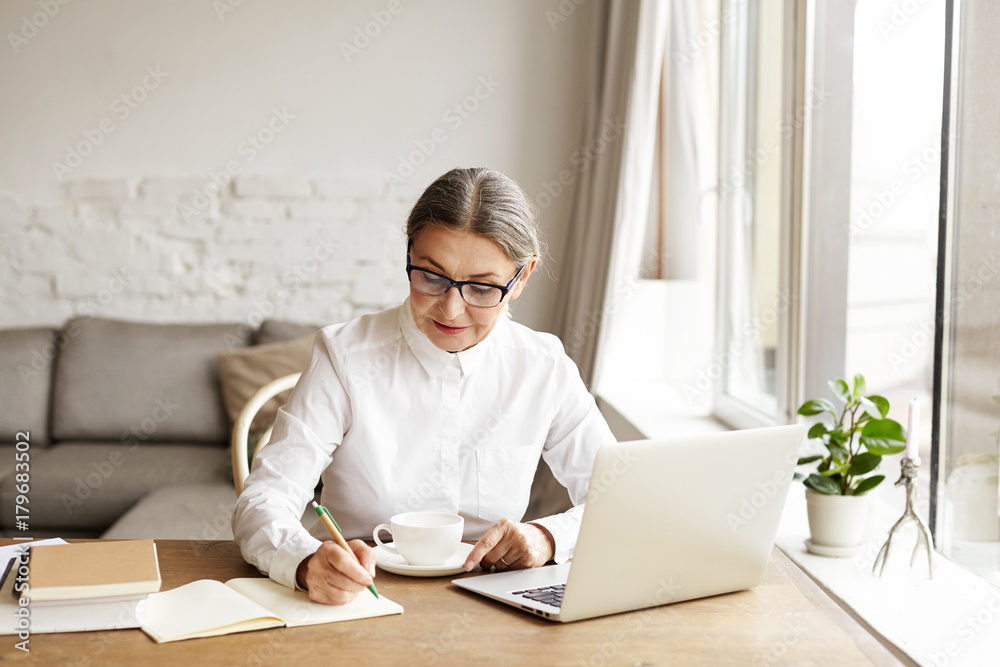 Picture of attractive mature woman business coach wearing white blouse and eyeglasses making notes in notebook while sitting at her cozy office, working on generic laptop computer and drinking coffee