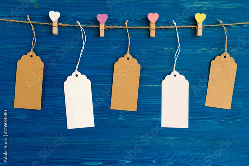 Brown and white blank paper price tags or labels set and wooden pins decorated on colored hearts hanging on a rope on the blue wooden background.