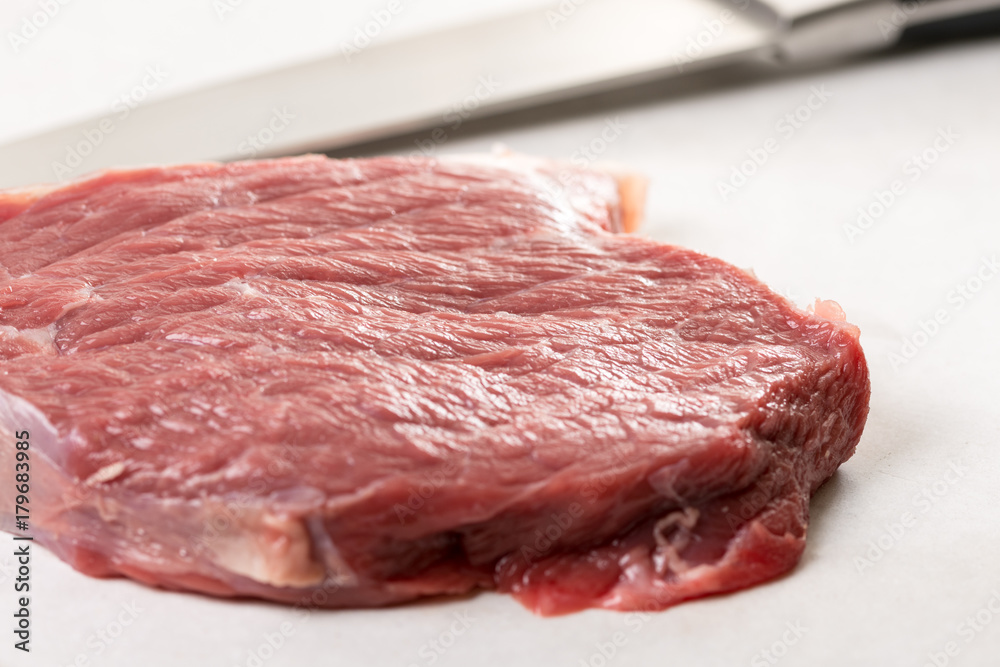 Raw beef steak on the white marble table with chef knife in the background Raw beef steak on the white marble table with chef knife in the background