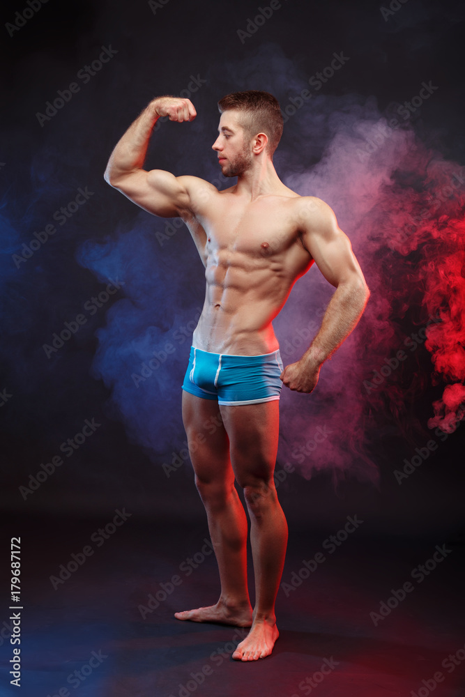 Young muscular ripped man bodybuilder with perfect body flexing muscles over dramatic smoky background. Power, strength, excellent body, bodybuilding, sports concept