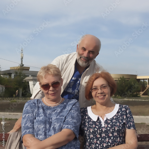 Two mature women sit on a bench, behind them a man stand. Warm September