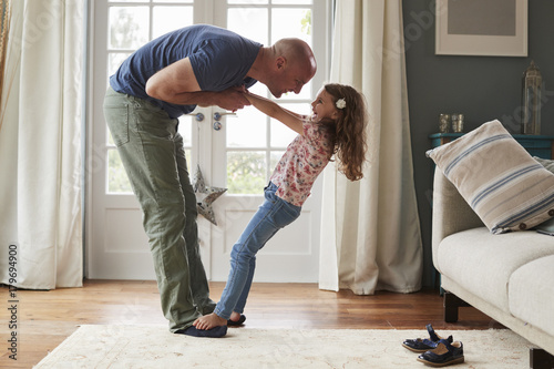 Girl balancing on father’s feet at home, side view