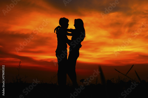  two people silhouetted against an orange sunset, embracing each other. They are standing in a field of grass.