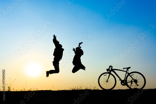  two people silhouetted against a sunset, jumping in the air next to a bicycle. © CreativeIMGIdeas