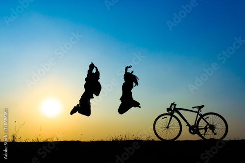 silhouette of young woman and boy jumping on sunset sky with bicycle on the prairie at yellow evening horizon sea yellow sunset heaven background Outdoor.