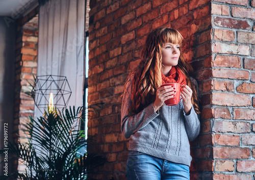 A woman drinks hot coffee near the wall of a red brick.