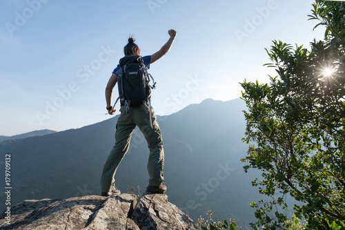 young woman backpacker standing on cliff's edge with raised hands