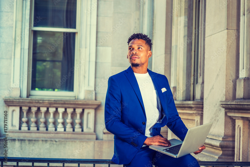 Foto de African American Businessman working in New York. Wearing blue suit,  unbuttoned, white T shirt, black college student with little goatee,  standing by vintage library doorway on campus, looking forward do