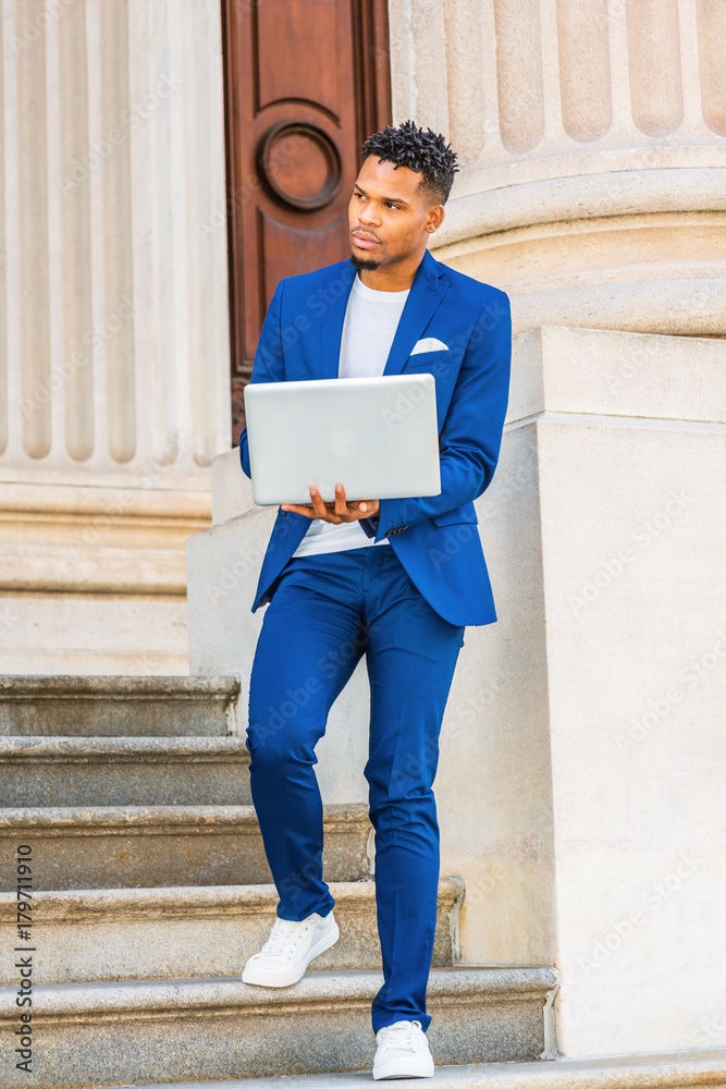 African American college student studying in New York, wearing blue suit, white  sneakers, standing on stairs of vintage office building on campus, working  on laptop computer, looking away, thinking.. Stock Photo
