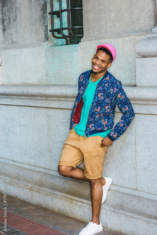 African American Man Casual Street Fashion in New York, wearing blue flower  patterned jacket, green V neck T shirt, yellow brown shorts, white sneakers,  pink cap, standing by vintage wall, smiling.. Stock
