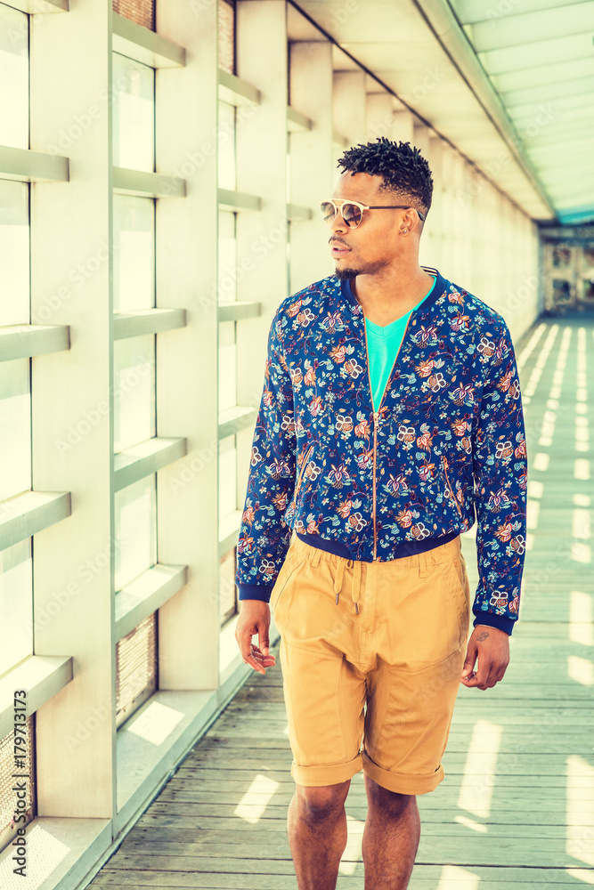African American Young Man Casual Fashion in New York, wearing blue flower patterned, collarless jacket, yellow brown pants, sunglasses, walking on walkway with glass wall on campus under sun..