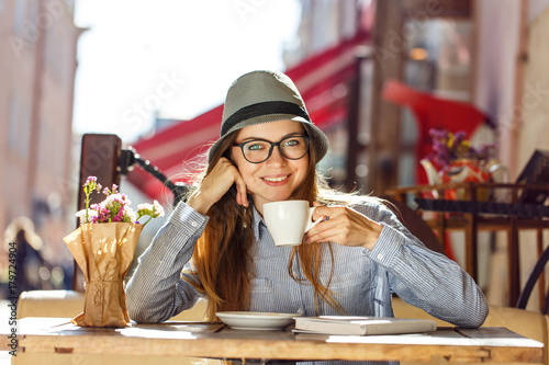 Stylish cheerful girl wears blue shirt, hat and eyeglasses having cup of coffee in street cafe, outdoors sunny autumn time