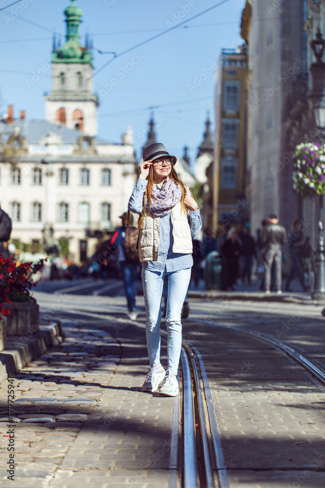 Casual stylish girl wears hat and eyeglasses walking in old city street, autumn sunny day