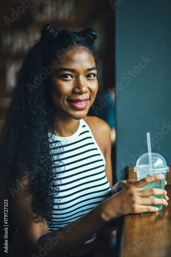 Beautiful smiling hipster teenager black ecuadorian woman tasting smoothie or juice in modern interior cafe. Relaxing, lifestyle and leisure concept. Fancy hairstyle like a cat. Close up. photo