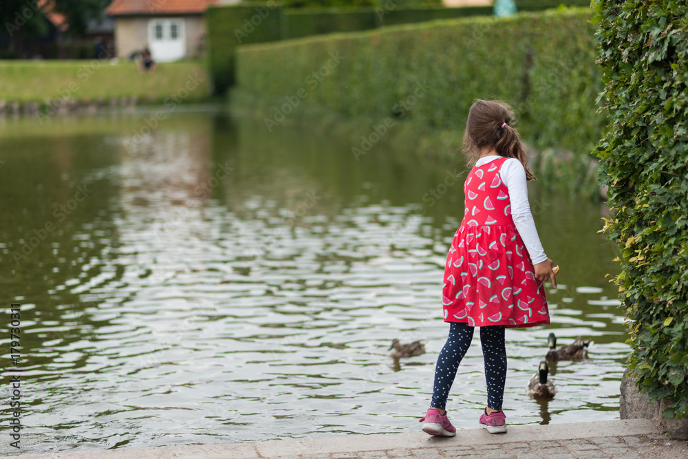 Young girls feeding birds by the city pond. Throwing bread to ducks. Pretty colorful dresses and skirts, long hair.