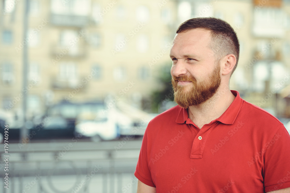man with a red beard, dressed in a red gaiety in the city. Happy and healthy. looks and mysteriously smiles. Concept idea of success man with a red beard, dressed in a red gaiety in the city. Happy and healthy. looks and mysteriously smiles. Concept idea of success