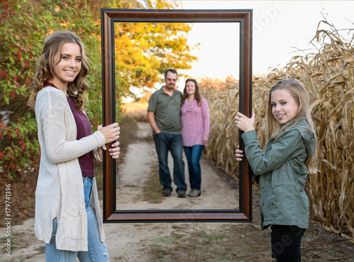 smiling blond sisters holding picture frame with parents blurred in background on rural dirt road