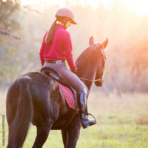 Rear view of teenage girl riding horse in park at sunset. Young rider girl on bay horse in the autumn park