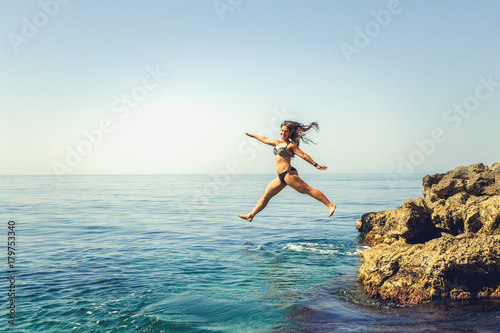 Cliff Jumping into the Ocean at Sunset, Summer Fun Lifestyle.Young girl in bikini joyfully free jumping off a cliff in to water.Woman jumping in blue water in tropical sea water on vacation.