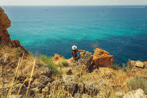 Young beautiful girl in a hat sits with her back to the camera on a turquoise summer background and enjoying the holiday of the soul.Rocky coast.Crimea.Fiolent.Clear water.One lonely boat in ocean.