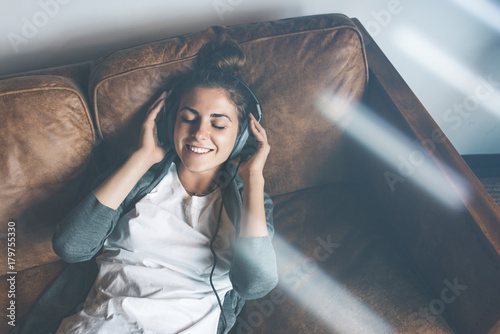Attractive young happy girl listening to music from headphones with pleasure at coworking office, lying on sofa and resting.