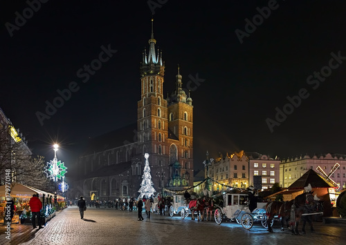 Christmas market and row of horse carriages on the Main Square of Krakow in front of the St. Mary's Basilica in night, Poland
