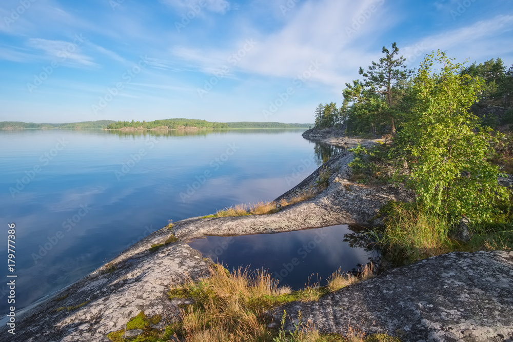 Landscape on the beautiful stone shore of the lake with islands in the summer morning with a beautiful sky and reflections. Ladoga Lake, Karelia, the island of Koyonsaari
