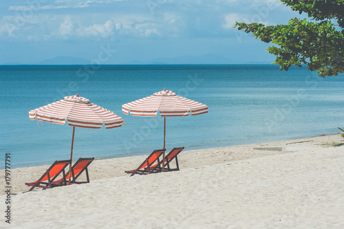 Vacation Concept   Four red wooden chairs and two beach umbrella setting on white sand with seascape and blue sky in the background.